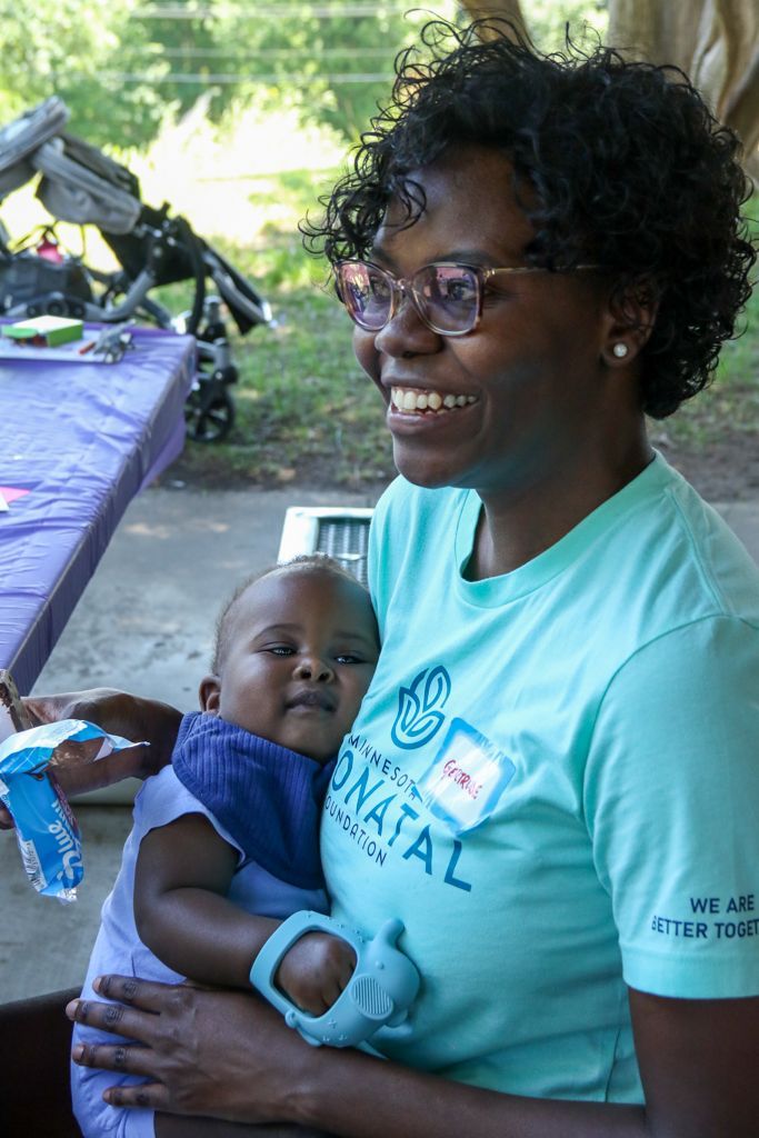 Woman smiling, holding baby. They are outdoors, baby wearing a bib, both with light blue accents.