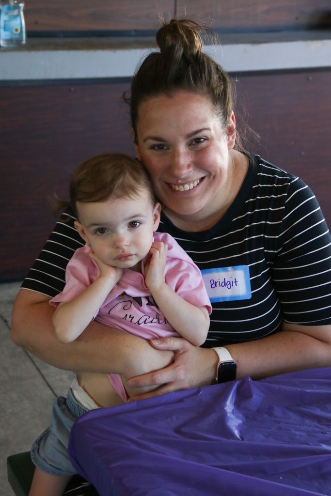 Woman smiling, holding child. Woman wears striped shirt, child a pink tee. Purple tablecloth.