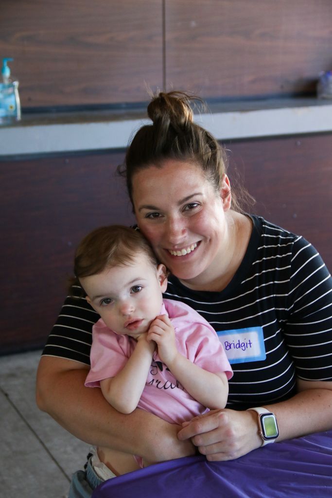 Woman with a bun and striped shirt holds a baby in a pink shirt; both smile. Inside, near a counter.