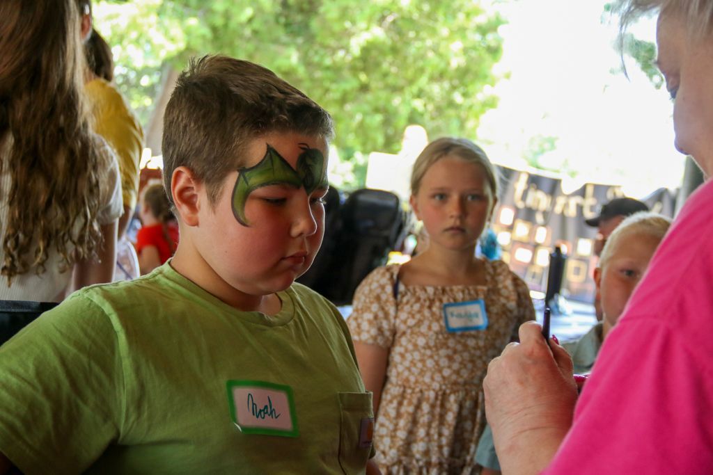 Boy with face paint, stands with a girl and a person with a pink shirt in front of him. Outdoors.