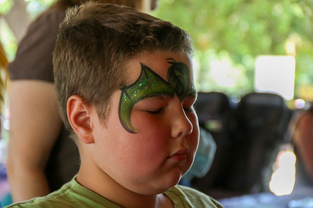 Boy with dragon face paint, eyes closed, cheeks blushed, outdoors.