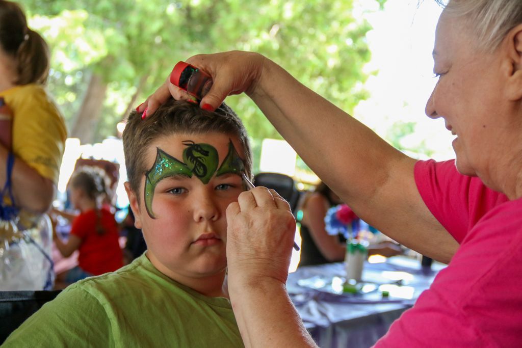 A person applying face paint to a boy outdoors. Green dragon design with an 