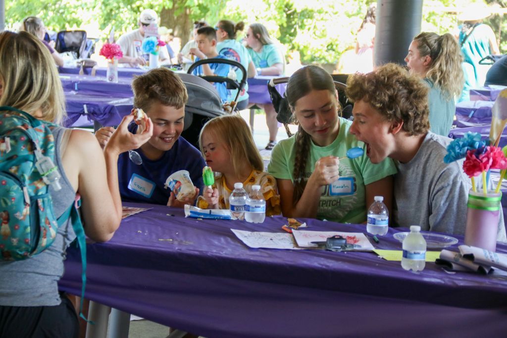People smiling around a purple table, possibly at an outdoor event.  Bottled water and decorations present.