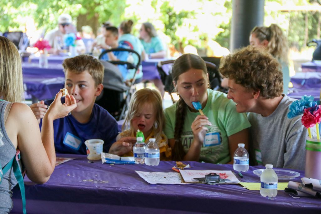 People at a table, eating lollipops and drinks outdoors under a canopy, with a purple tablecloth.