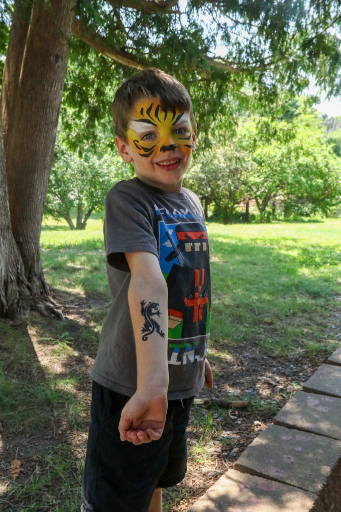 Boy with tiger face paint and a tattoo, standing outdoors, smiling.