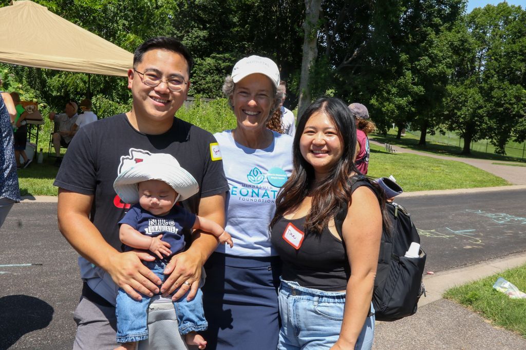 Group of four people smiling outdoors: man holding baby, woman with hat, young woman, with green trees in background.