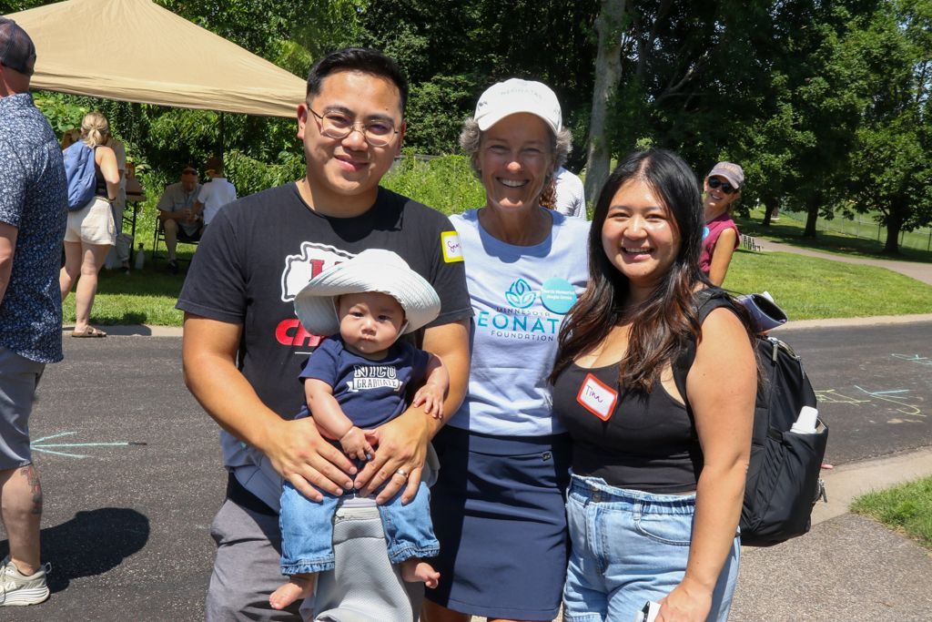 Family poses outdoors: man holding baby, woman in a hat, and another woman smiling. Green background.