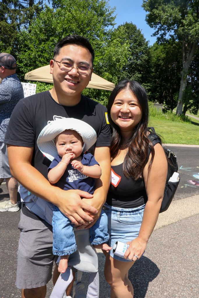 Family of three smiles outdoors: dad holds baby, mom stands beside. Sunny day, trees in background.