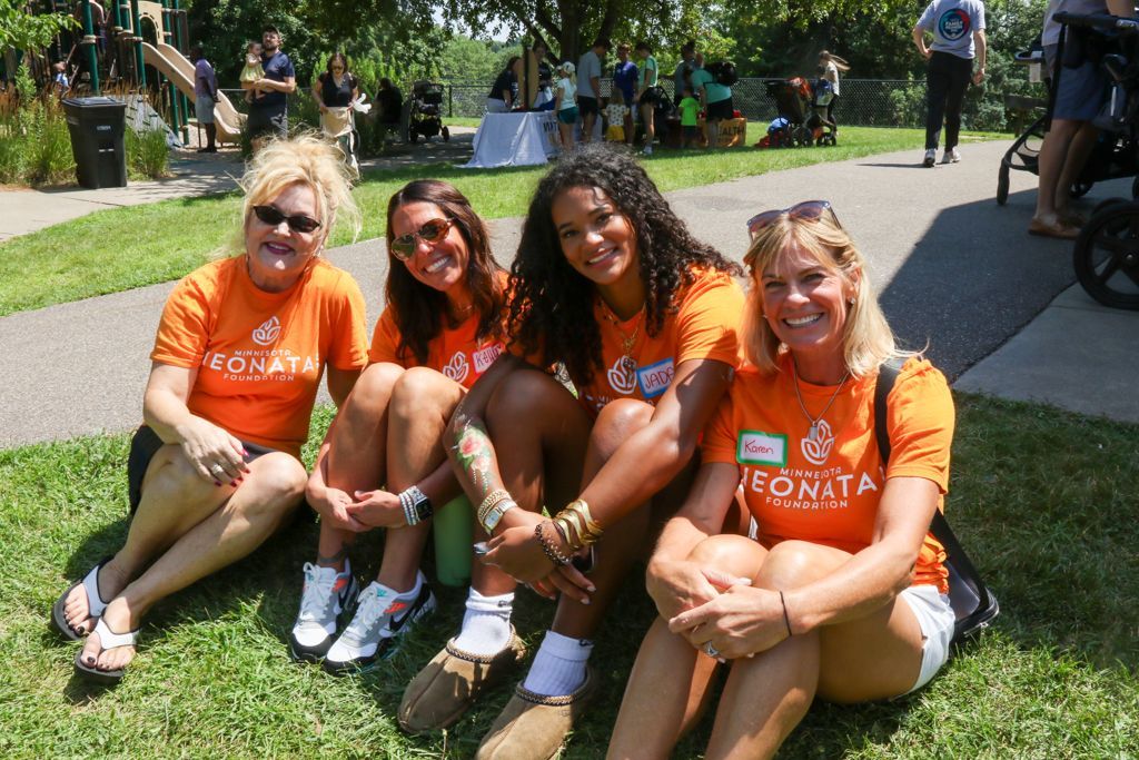 Four people in orange shirts sit on grass, smiling. Outdoor event setting.