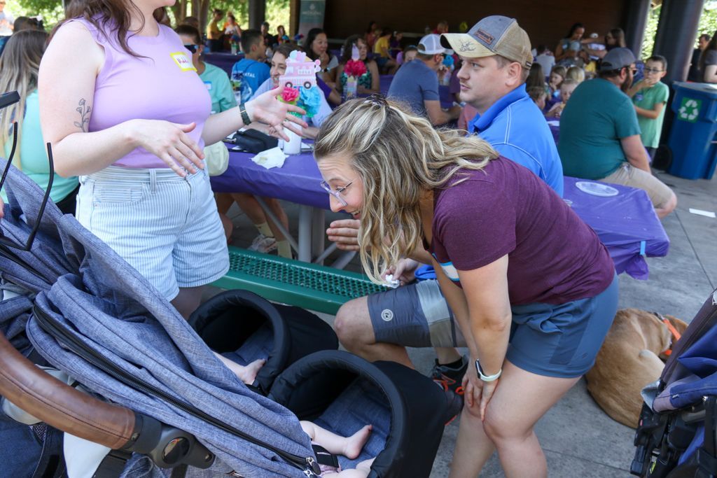 People at an outdoor event. Woman in maroon top leans toward a stroller. Others seated at a purple table.