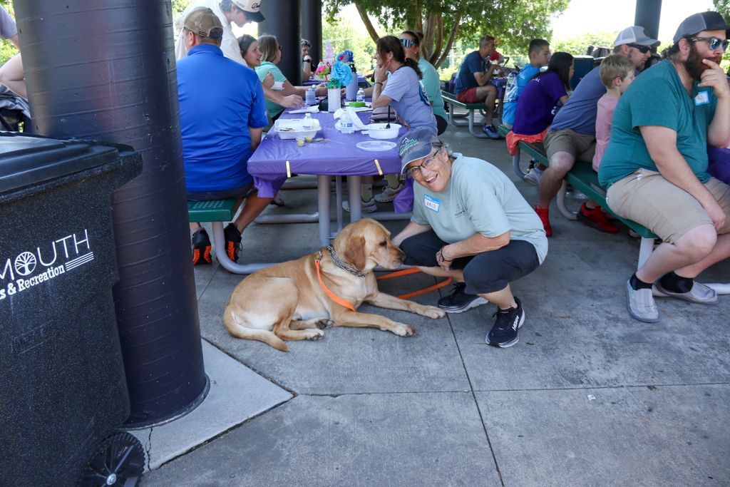 A woman pets a golden lab on a leash at a picnic table, other people sit nearby, outdoor setting.