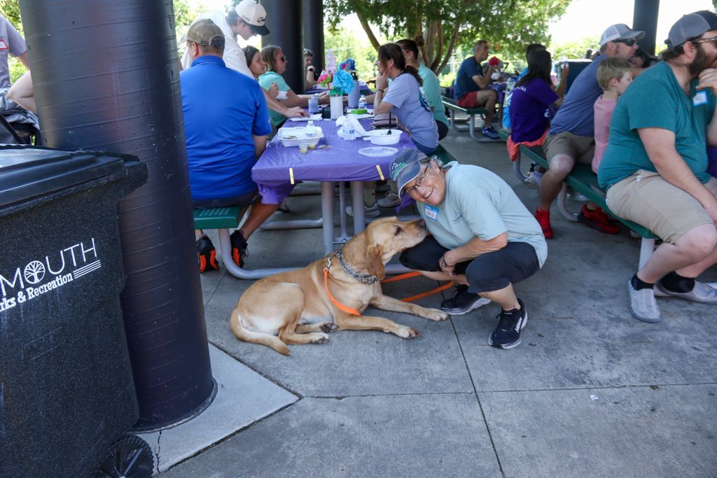 Woman petting a golden Labrador on a leash; people seated at picnic tables in a park.