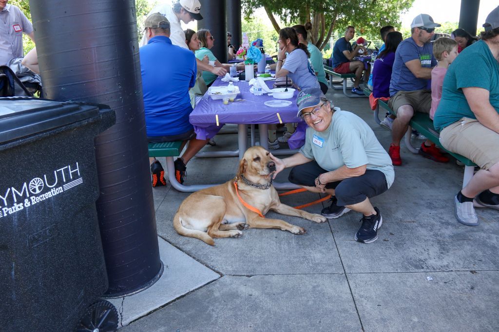 A person in a teal shirt kneels beside a yellow lab on a leash in a park; people seated at picnic tables.