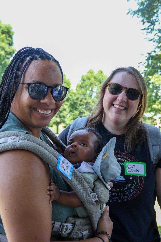 Two women and baby in carrier outdoors, smiling. Green trees in background.