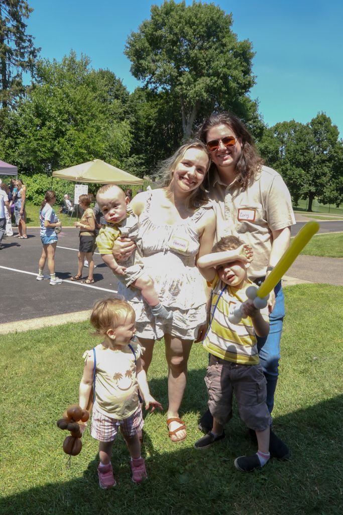 Family of five smiles outside on a sunny day. Two adults, three children. One child holds a yellow bat.
