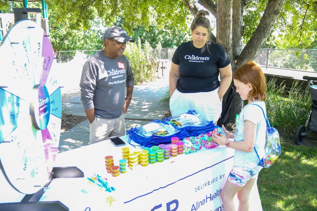 Two people at a table assist a child, possibly at an event. Colorful prizes and a spinning wheel are visible.