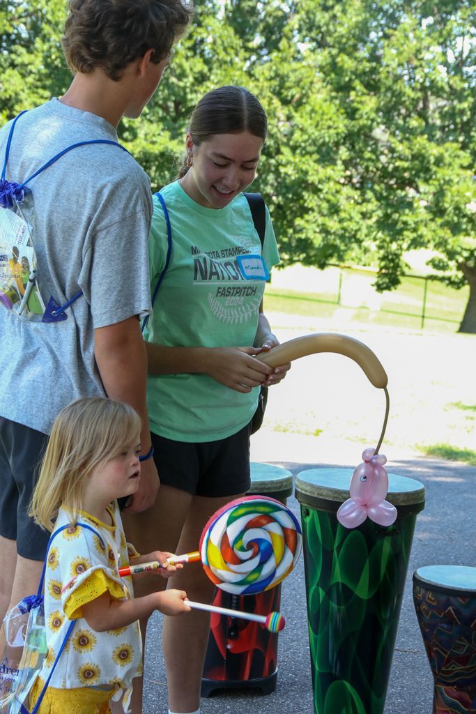 Three people with balloons and a lollipop by decorated drums outdoors.