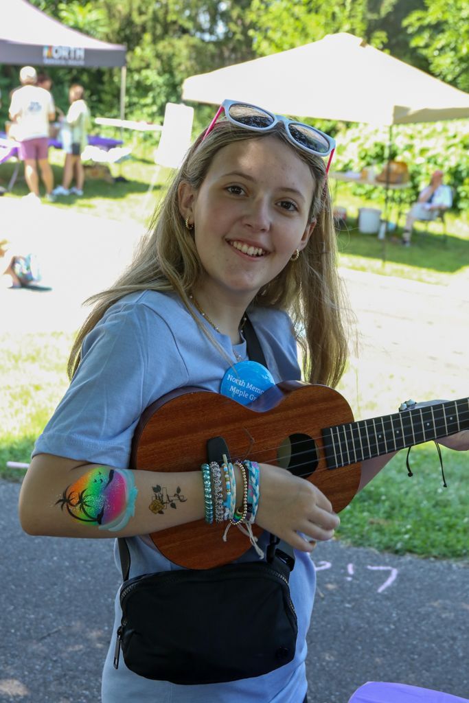 Girl smiles while playing ukulele outdoors, wearing sunglasses on head, arm tattoos, and multiple bracelets.