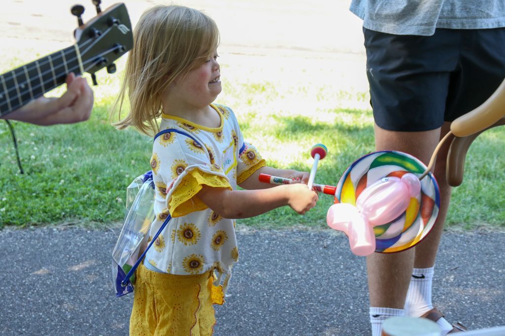 Child playing a toy with colorful spinning parts, smiling outside. Person plays ukulele.