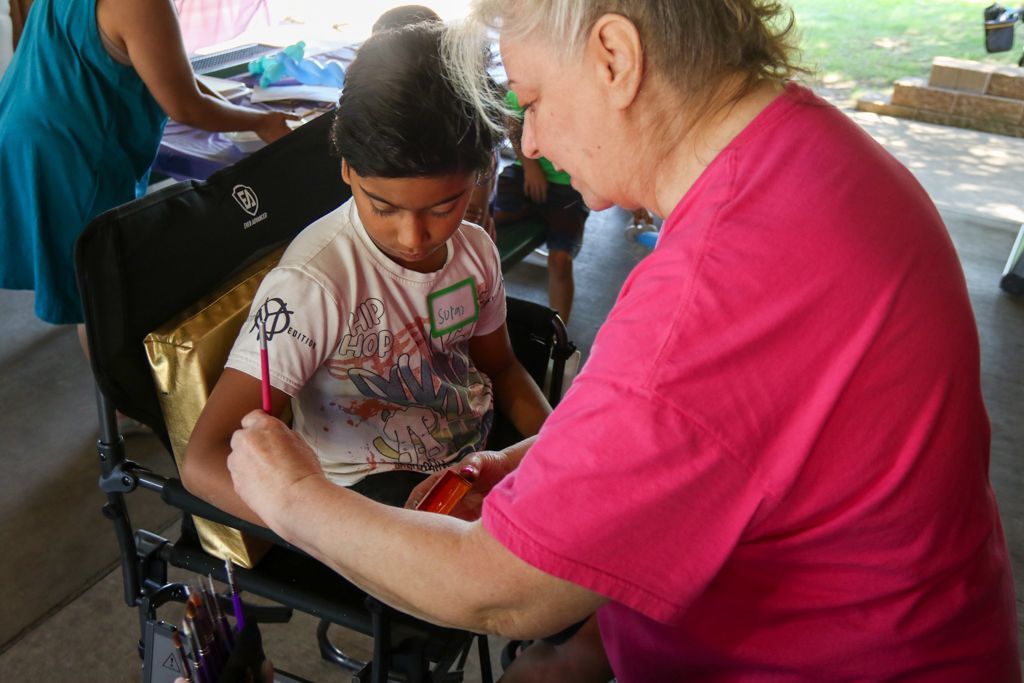 Woman painting on boy's arm outdoors. Boy sits in a chair, wearing a turban and shirt with paint on it.