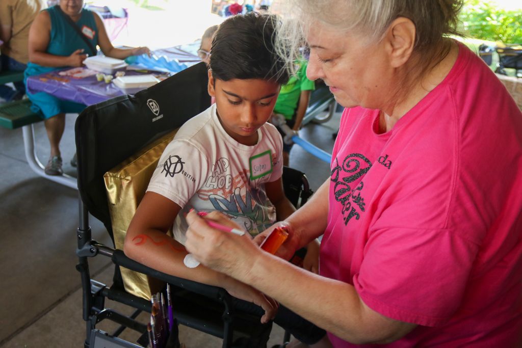 Woman paints a design on a child's arm outdoors. Child sits in a chair, focused.