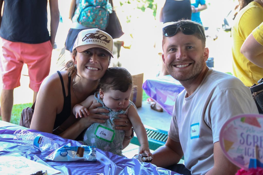Family at an outdoor event, smiling. Woman holding baby, man at right. Purple tablecloth, sunny day.