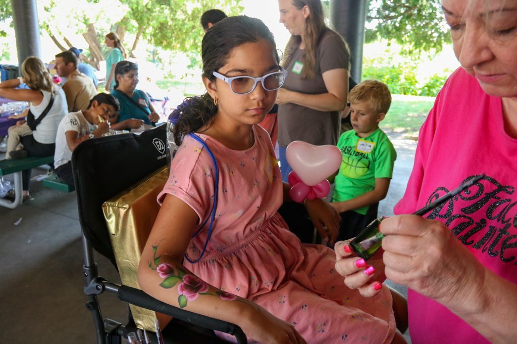 Girl getting arm painted with flowers at an outdoor event; woman paints. Other people in the background.