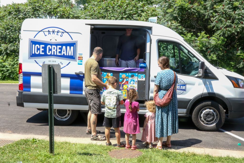 Ice cream van with customers: children selecting treats with adult supervision. Van is white with blue accents.