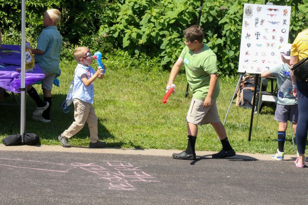 Two boys play with toy guns on a paved area, green trees in background, display board nearby.