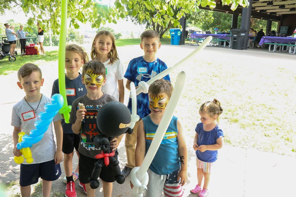 Group of children holding balloon swords, some with painted faces, at an outdoor event.