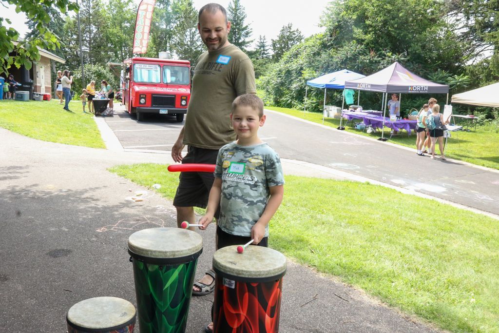 Man and boy play drums on a path near tents and a red truck.
