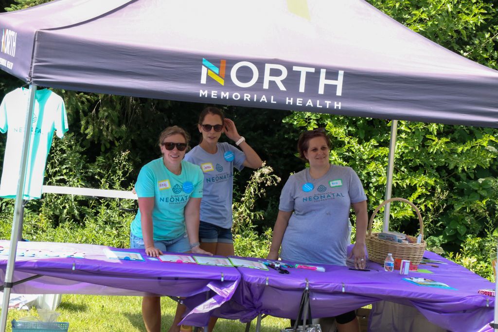 Three people at a North Memorial Health booth, under a canopy. They are wearing light blue shirts and smiling.