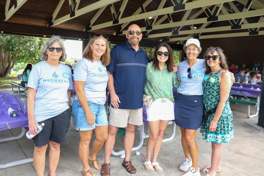 Group of people smiling outdoors under a gazebo, some wearing light blue shirts and sunglasses.