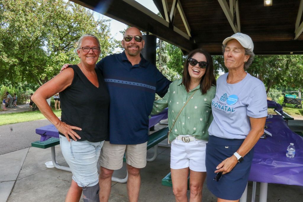 Four people smiling under a covered outdoor structure. Greenery in background, purple picnic tables.