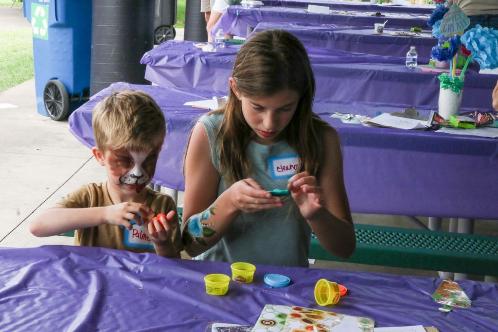 Two children at a crafts table, working with playdough. Boy has face paint. Purple tablecloth. Outdoor setting.