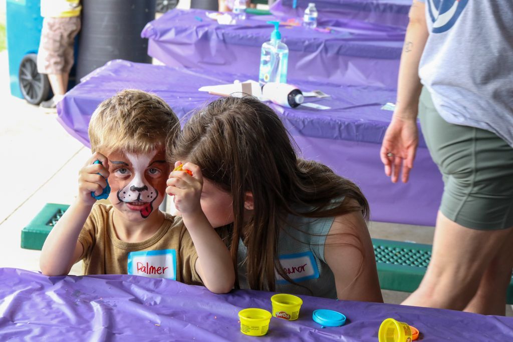 Two children at a table with play dough; one has face paint of a dog. Purple tablecloth.