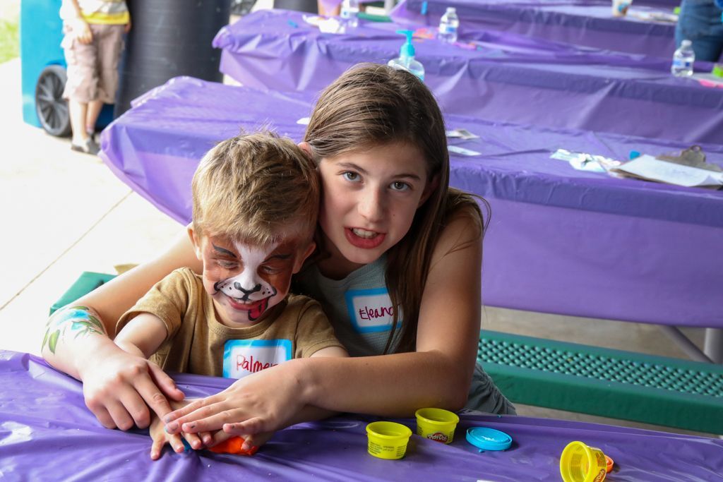 Girl with arm around boy with face paint at table with purple tablecloth.