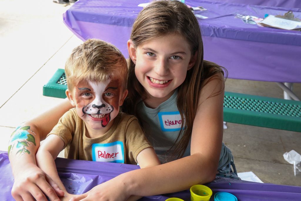 Girl and boy at a table, both smiling; boy has face paint, girl's arm around him. Purple tablecloth.