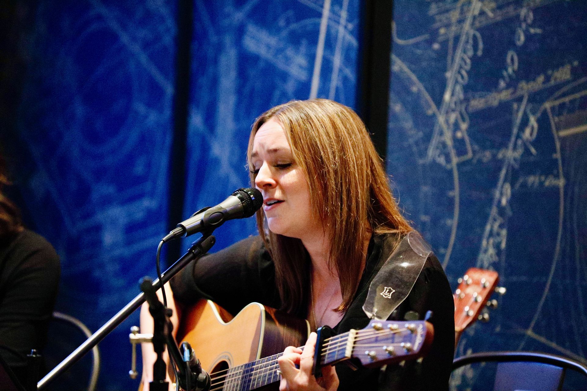 Woman playing acoustic guitar, singing into microphone. Dark-haired, black shirt, performing. Blue backdrop.