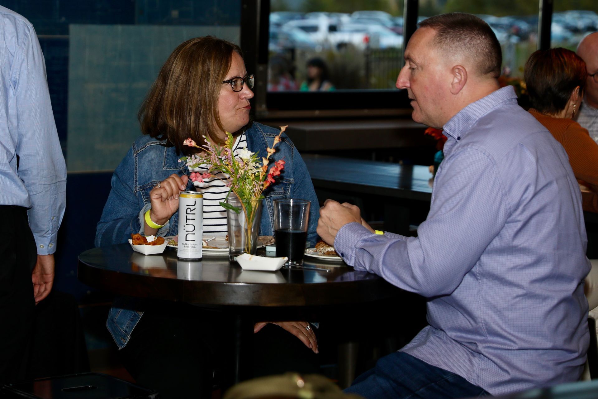 Two people seated at a round table in a bar, chatting and eating snacks.