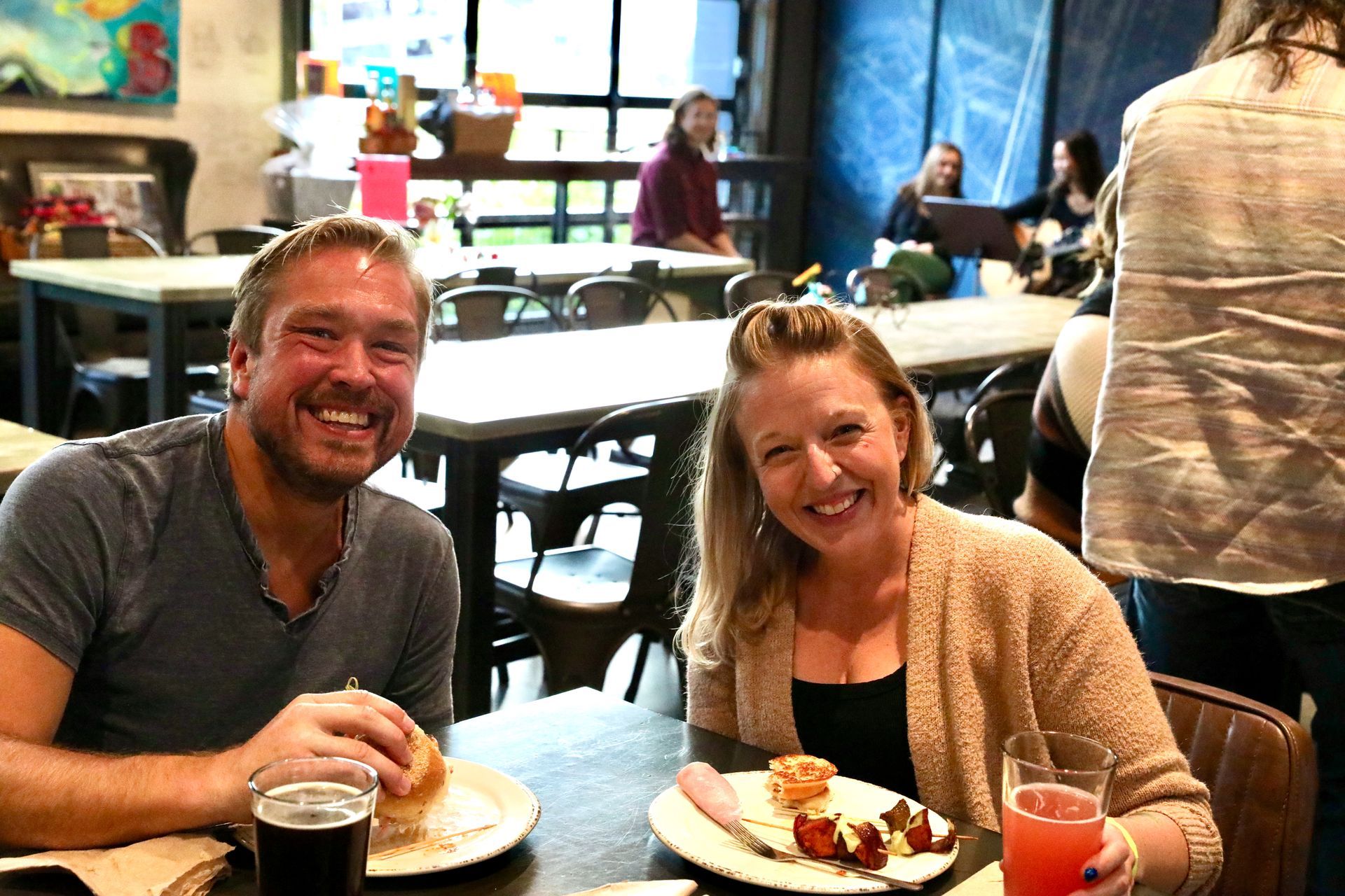 Man and woman smiling at a table, eating and drinking in a restaurant. Others in background.