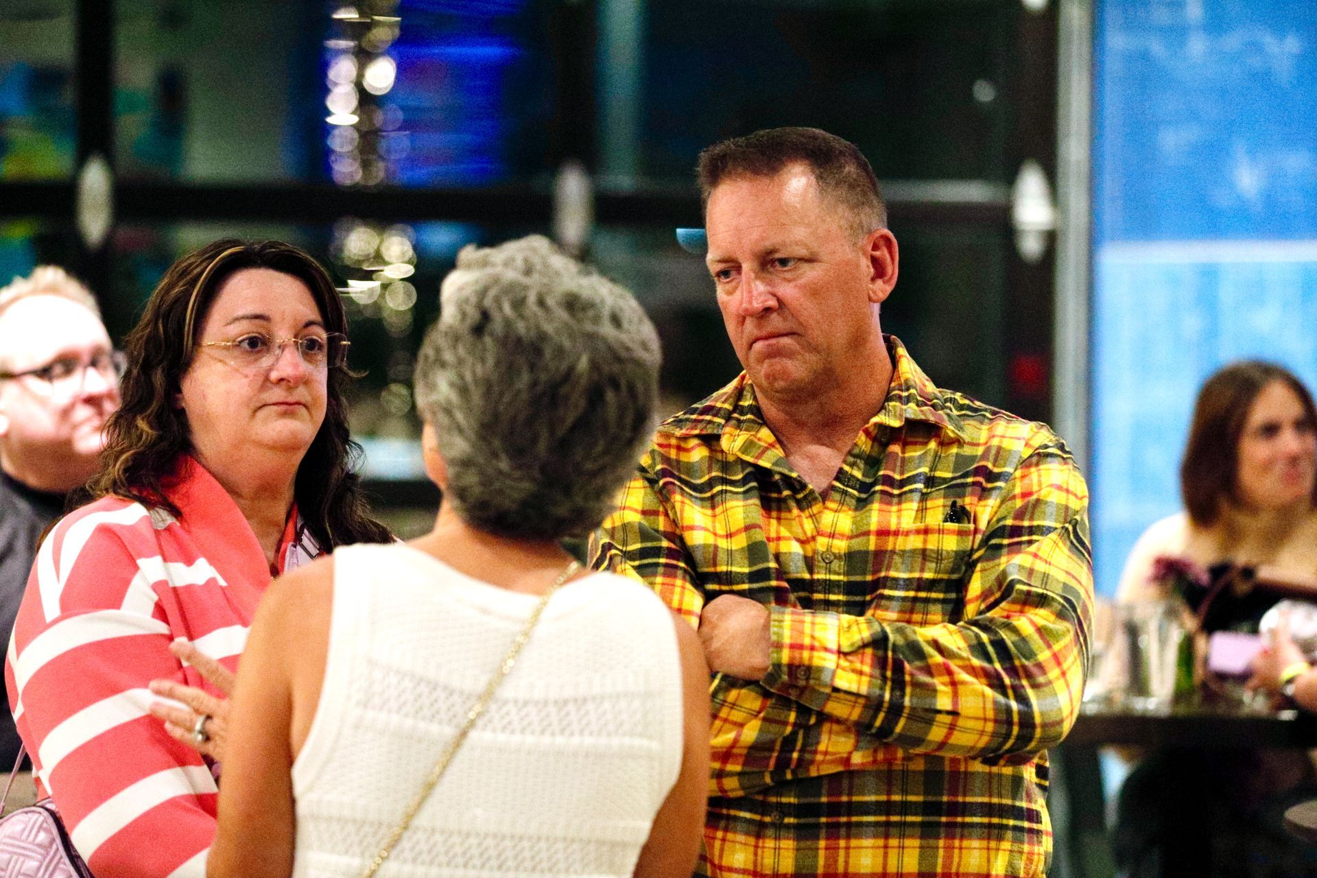 People in a crowded room, man in yellow plaid shirt arms crossed, woman in pink and white stripes nearby.