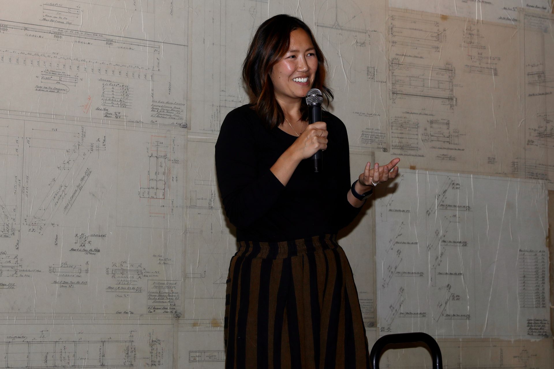 Woman with microphone speaks, gesturing, in front of a textured wall with writing.