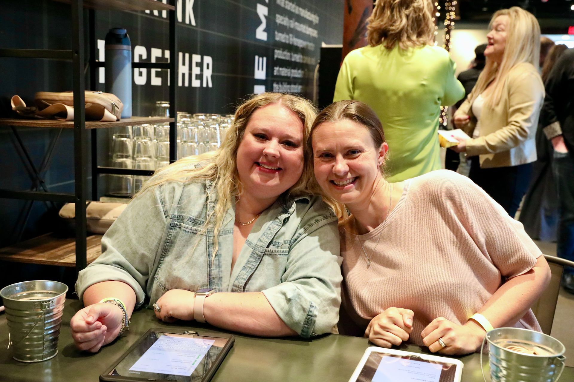 Two women smiling at a table, one in denim, one in pink, at a modern indoor event.