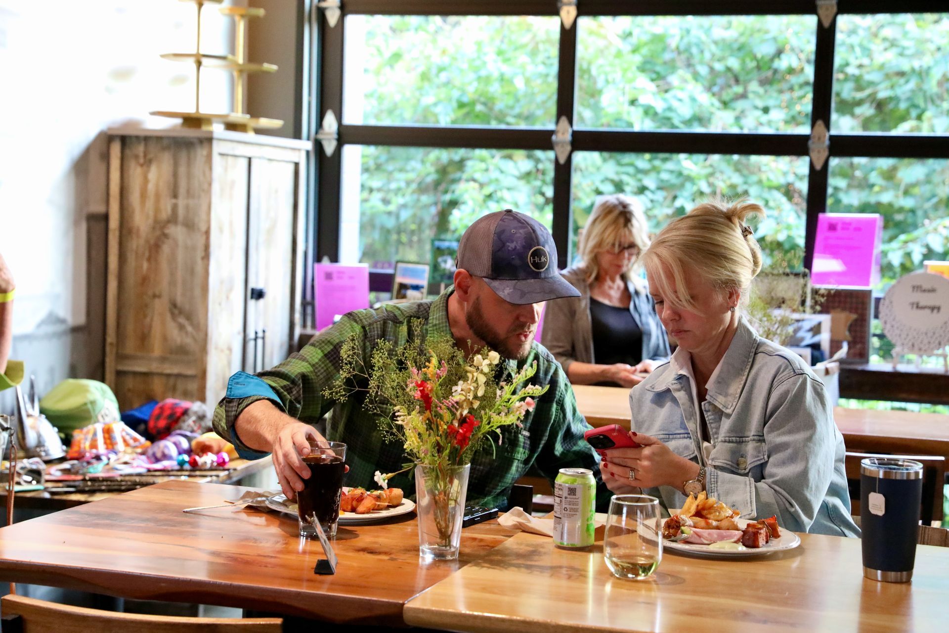 Two people at a table in a cafe, looking at a phone, food and drink present. Other people and a window in the background.