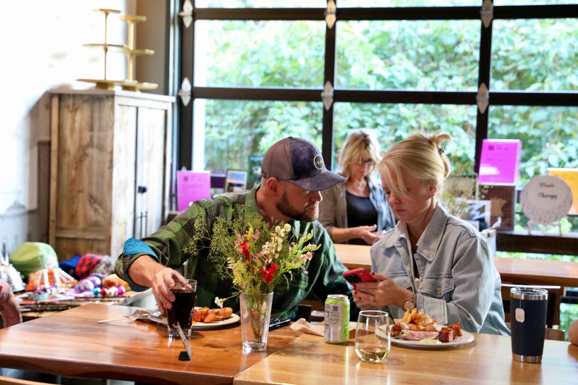 People at a restaurant table, one looking at phone, the other holding a drink, food and drink on table.