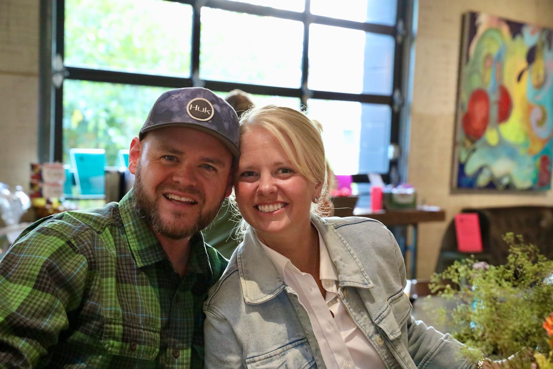 Man in green plaid shirt and woman in jean jacket smile, indoors, near a window.