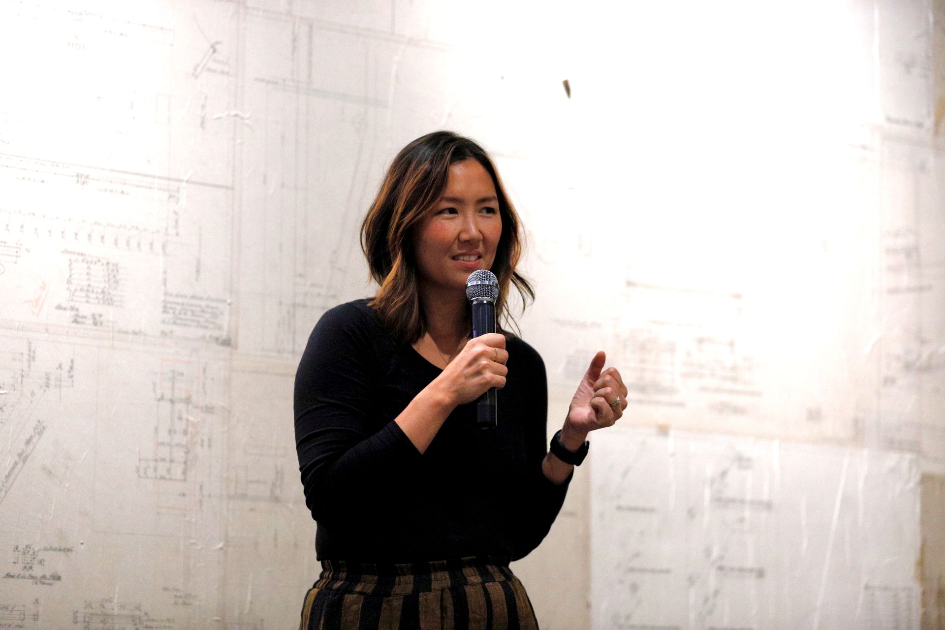 Woman speaks into a microphone, gesturing with her right hand. She wears a black shirt and striped pants, standing in front of a white wall with writing.