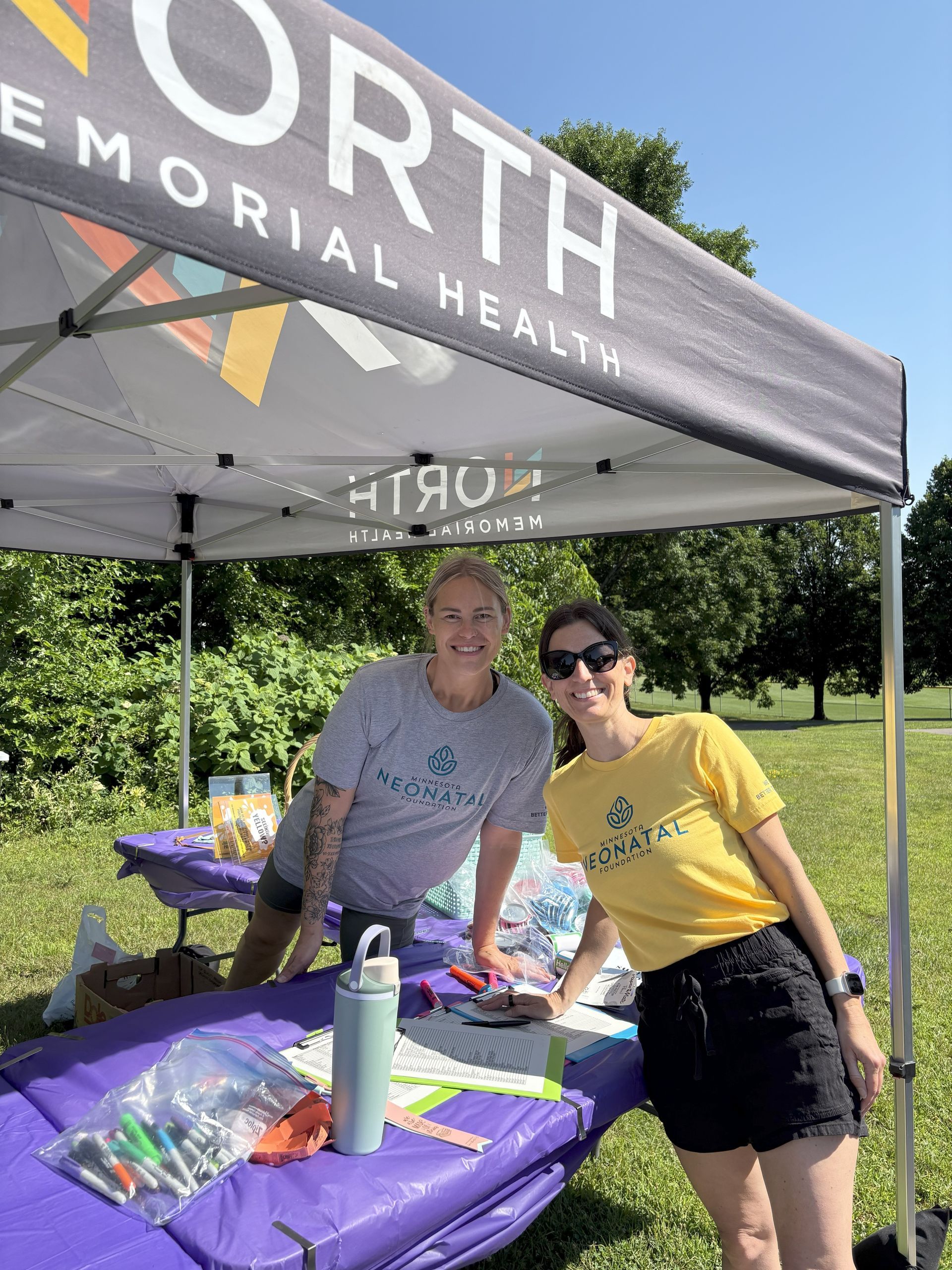 Two women smiling at a North Memorial Health booth in a park.