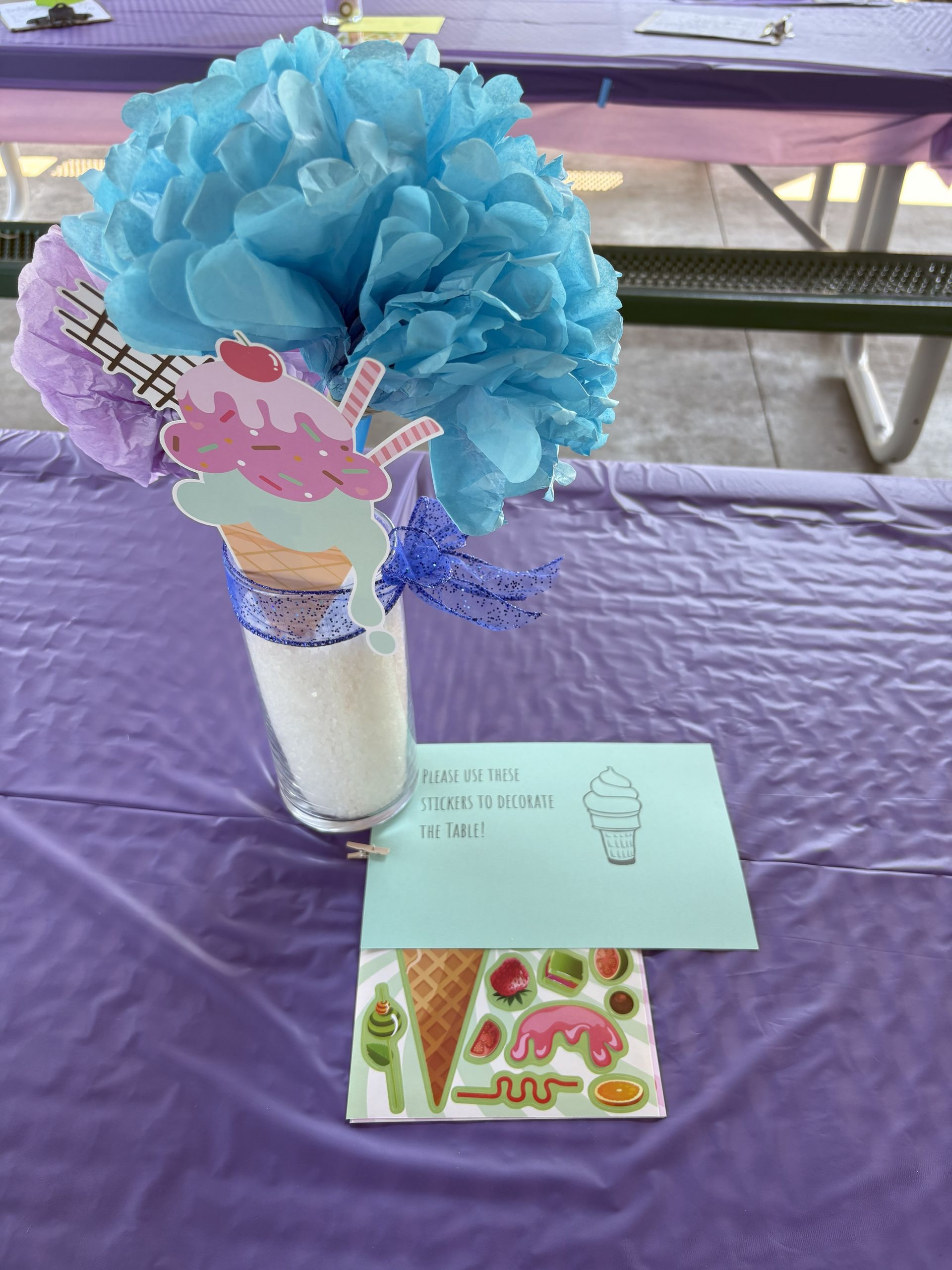 Ice cream cone centerpiece on purple tablecloth, with paper flowers and craft supplies.